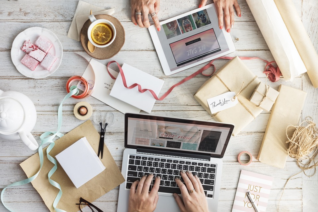 Two people working on a desk filled with gift wrapping objects on their laptop and tablet.