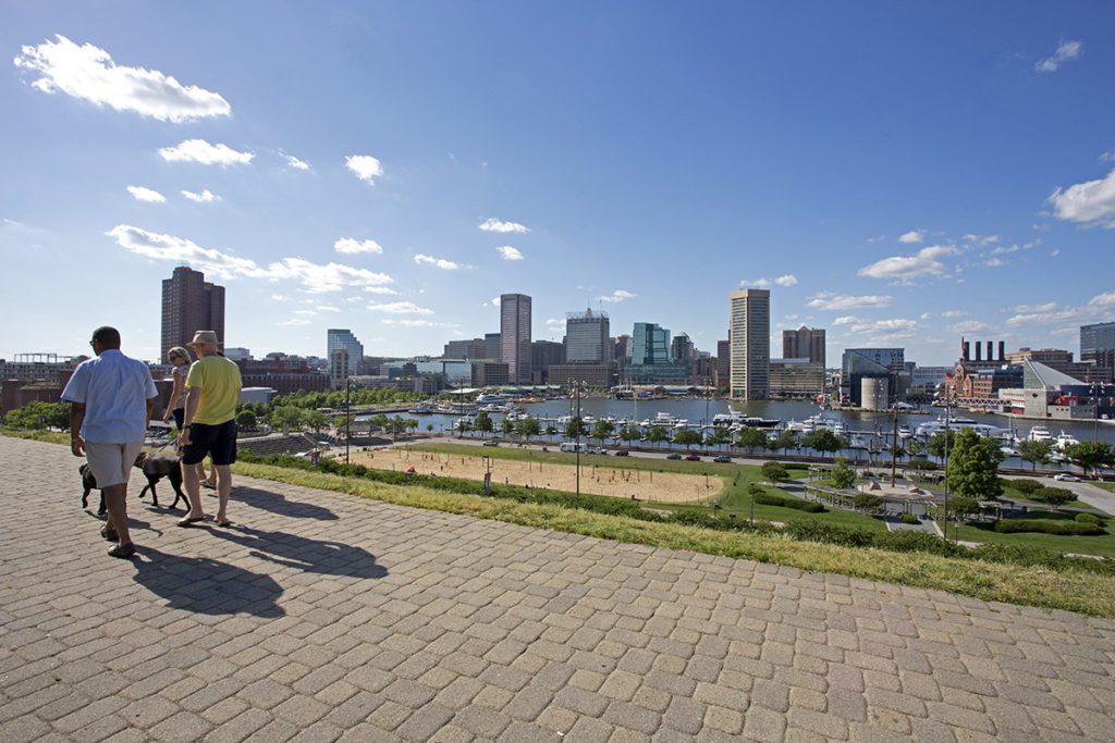 Three people walk together with two dogs along a pathway overlooking a park and Baltimore's waterfront.