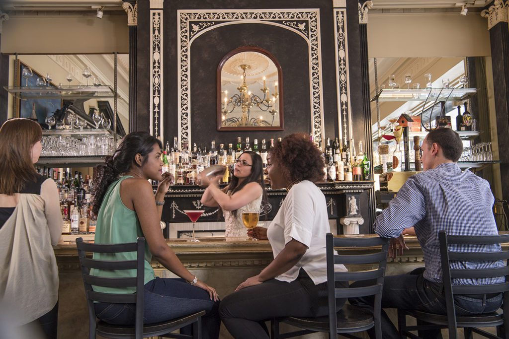 Four people sit at a bar with Edwardian architectural details, as a bartender mixes a cocktail in the background. Affordable event destinations benefit from low-cost living in multiple ways, including the ability to attract top talent in fields like the culinary arts and drink mixology.