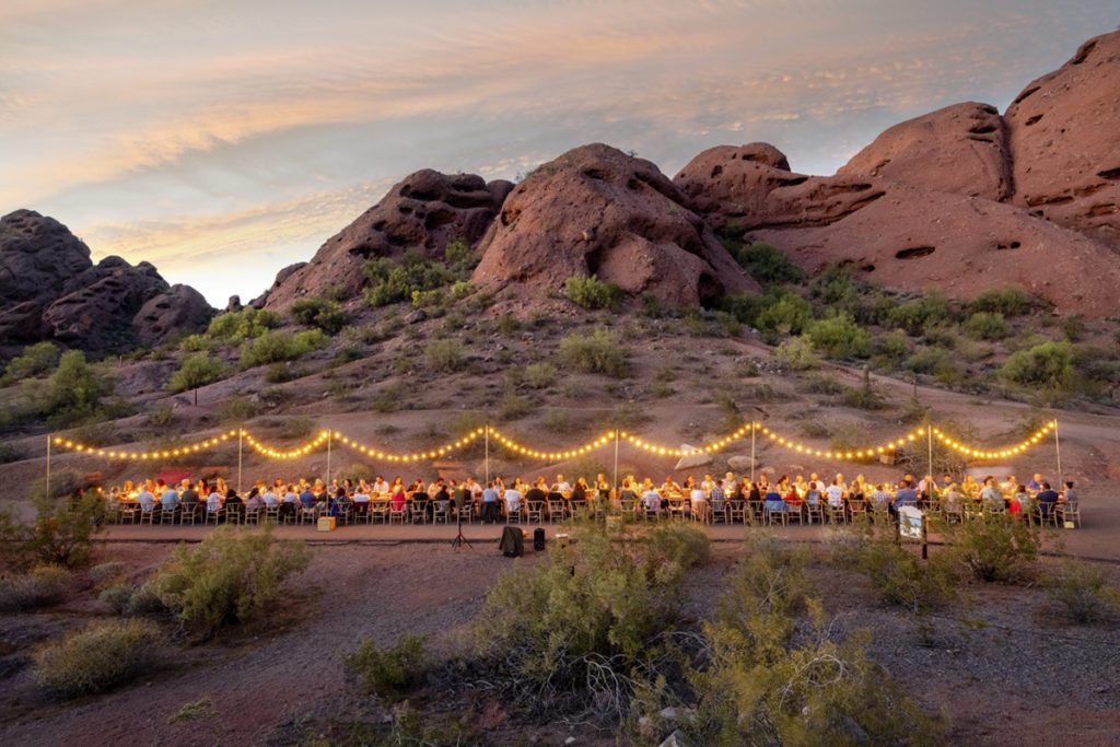 An evening scene of an outdoor banquet with around 100 people seated along on long table under string lights in Arizona's Sedona Desert, just outside Phoenix.