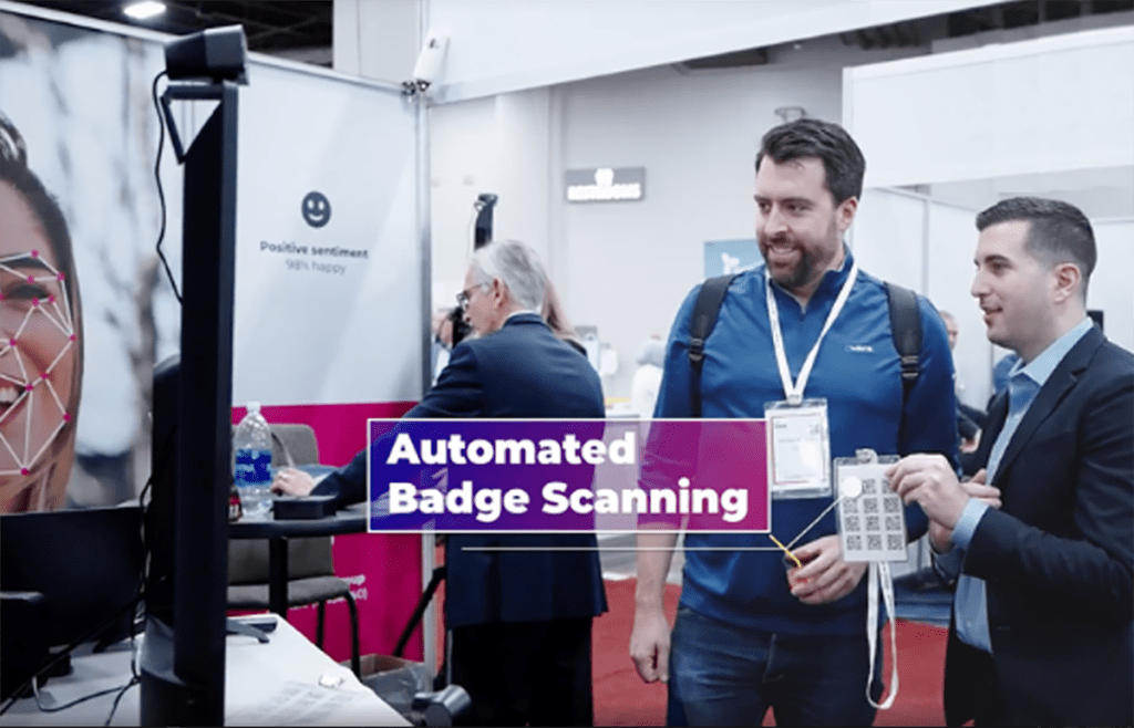 Two conference attendees demonstrate Zenus AI's automated badge scanning tool by standing in front of a dedicated camera that detects the QR code on their registrant badge.