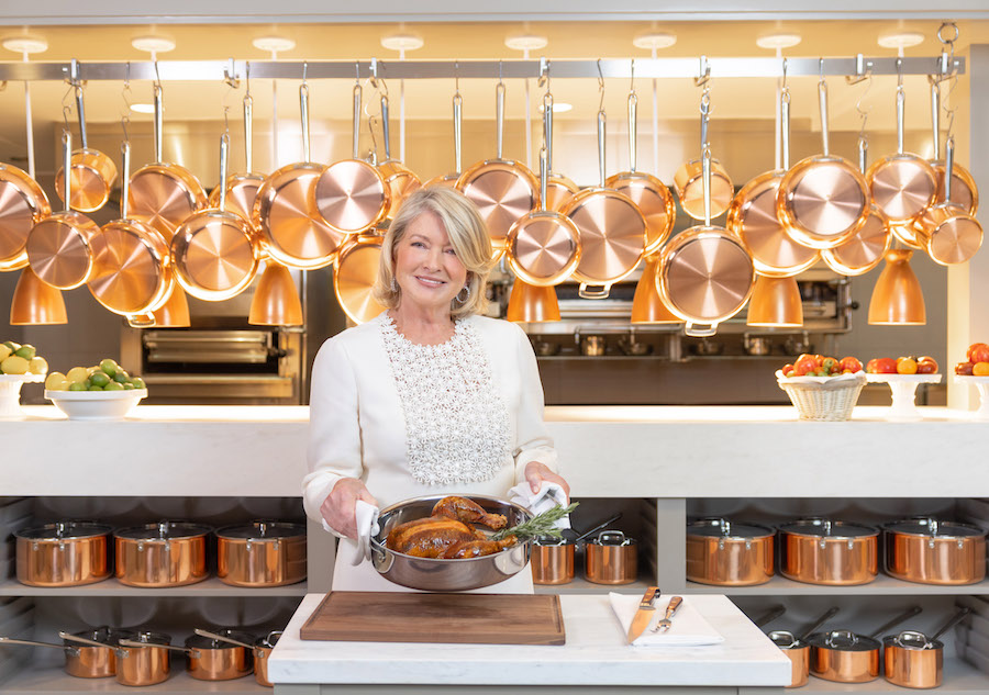 Martha Stewart stands holding out a roast chicken in front of a kitchen with multiple copper pans and pots on display at the Bedford restaurant in Las Vegas.