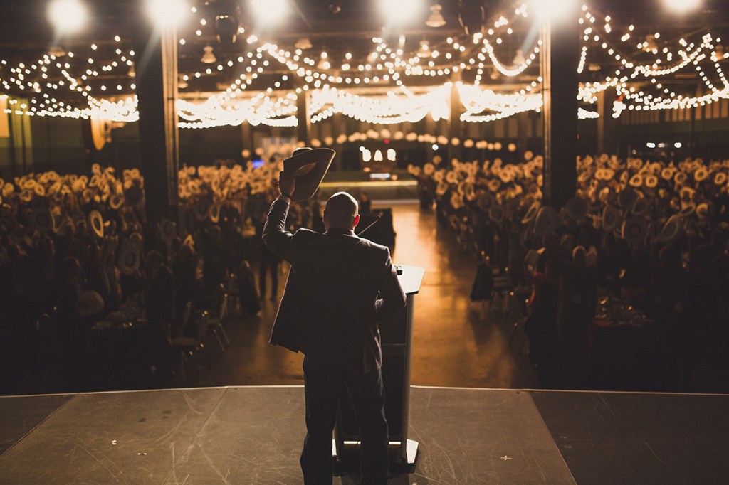 A view from the back of a stage looking out at the audience, with the speaker and the audience lifting white hats as part of Calgary's welcoming White Hat Ceremony — a key aspect of the city's culture of outstanding hospitality.