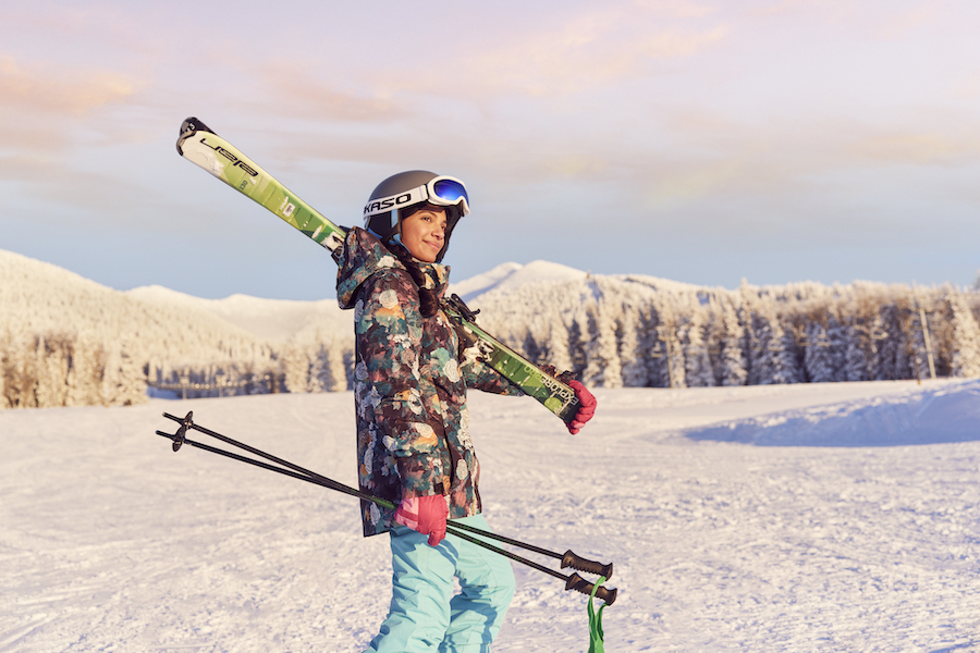 A woman stands holding skiing gear with a snowy backdrop of pine trees and mountains of Arizona (demonstration of the meeting myth that you can't have two seasons in one meeting).