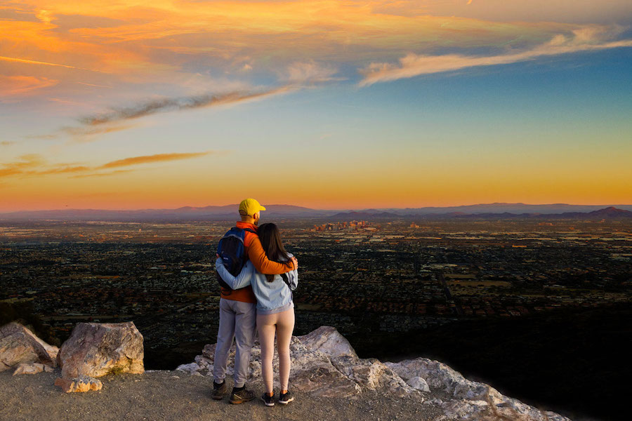 A man and a woman look out at Phoenix from a great distance while standing on a clifftop at sunset.