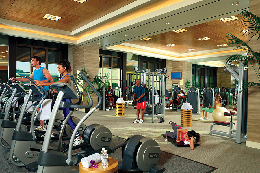 Two people work out on stepper machines in the foreground of a fitness center at Caesars Entertainment, with multiple other gym-goers visible in the background on different equipment that can help to maintain planner wellness.