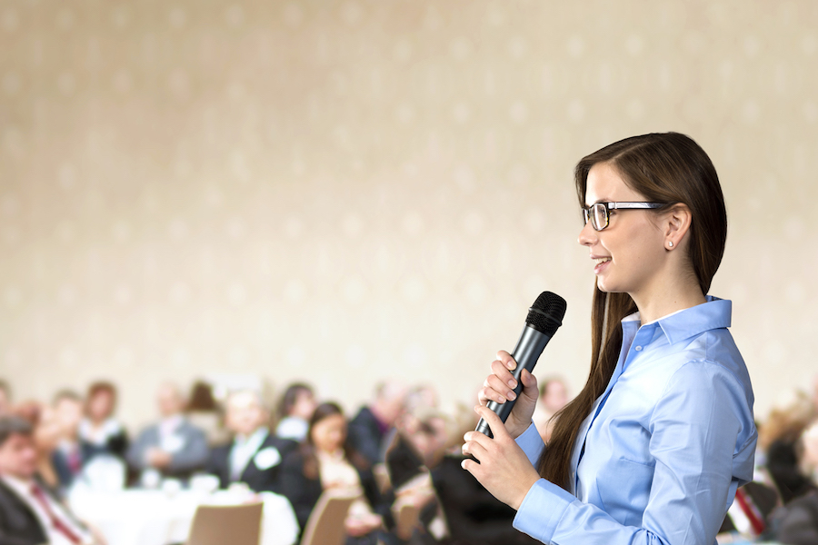 A business woman stands holding a microphone and speaking to a crowd of people inside a conference ballroom.