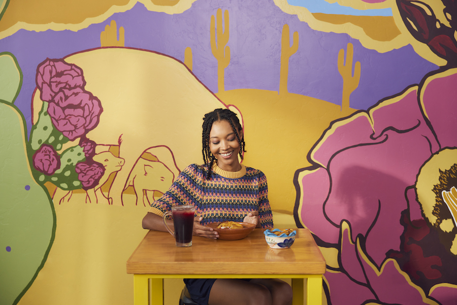 A woman smiles at her plate of nutritious food as she embraces the slow food movement's spirit of savoring F&B experiences while sitting in a restaurant covered in bright murals of local scenery.