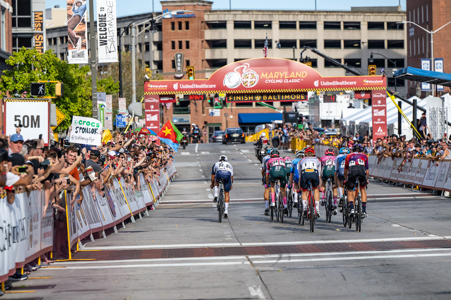 A view from behind of approximately ten cyclists as they approach the finish line at the Maryland Cycling Classic held in the classic sporting event host city, Baltimore, where plenty of fans are ready to cheer on the athletes.