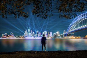 A man's silhouette stands at the waterfront of Sydney Harbour, with bright coordinated lights visible in the background as part of the Vivid Sydney event.