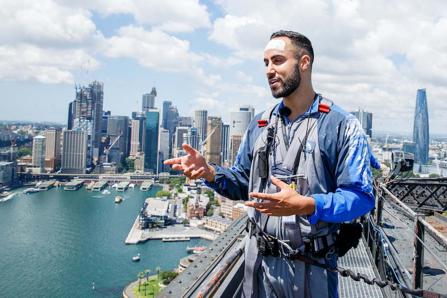 A First Nations storyteller stands in the foreground teaching a lesson on the history of Sydney to participants as part of an immersive bridge climbing activity organized by event changemakers.