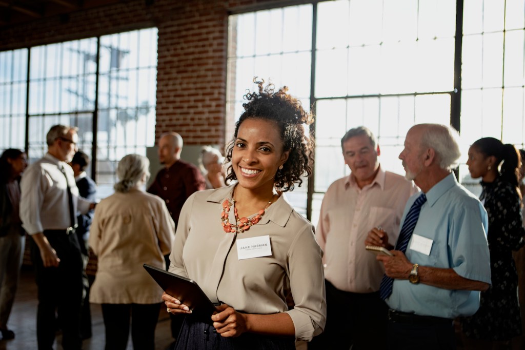 A business event marketer stands in the foreground of a networking activity, with approximately 10 attendees visible in the background.