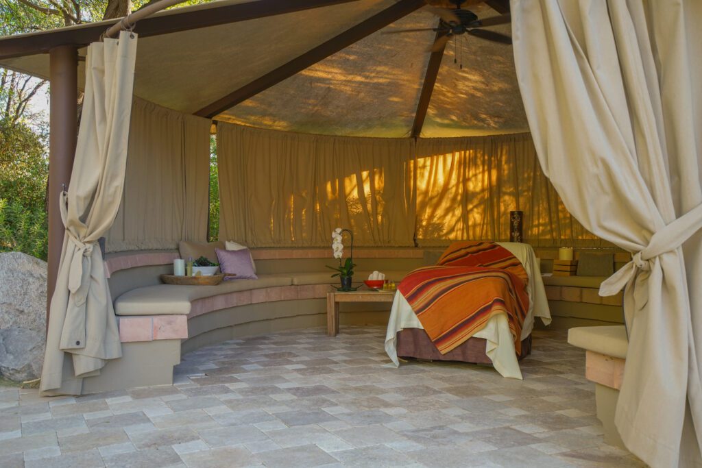 A large tented structure with wooden beams, a thick canvas covering, and a curved, semi-circle seating area at the base. A massage bed sits in the cent of the structure with an orchid, hand towels, and oils next to it — the perfect setting to fight digital burnout.