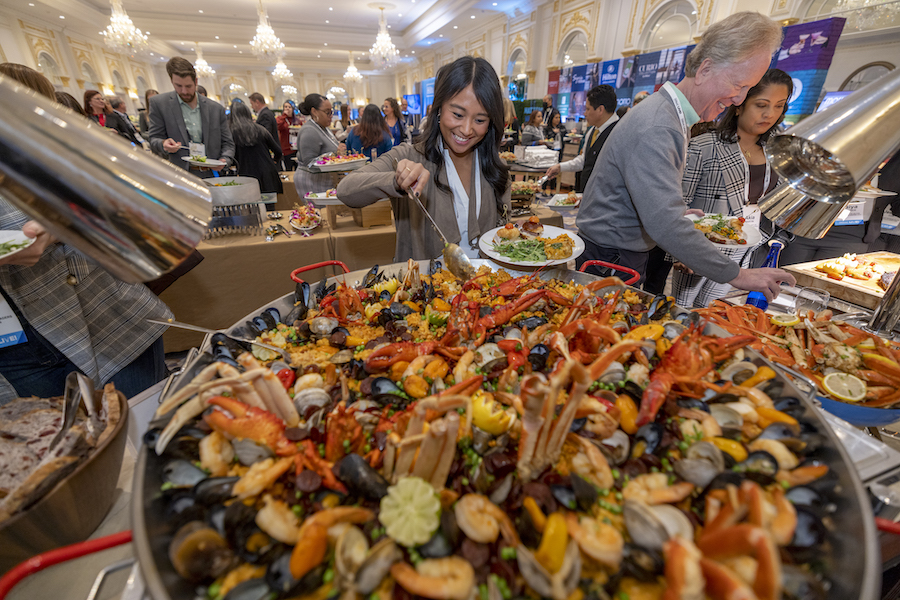 With two people beside her and multiple visible in the background smiling as they choose from row after row of a gourmet buffet, a young event professional spoons from an extra-large bowl of paella packed with mussels, lobster, and crab legs — with great food being one of the industry's top job perks.