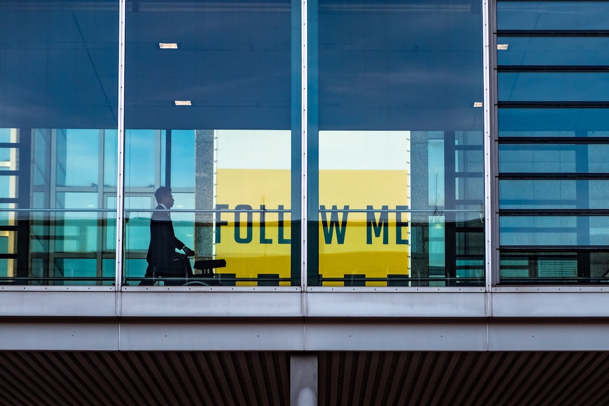 Business man in a suite and tie walking in an airport with a large yellow sign saying follow me in the background