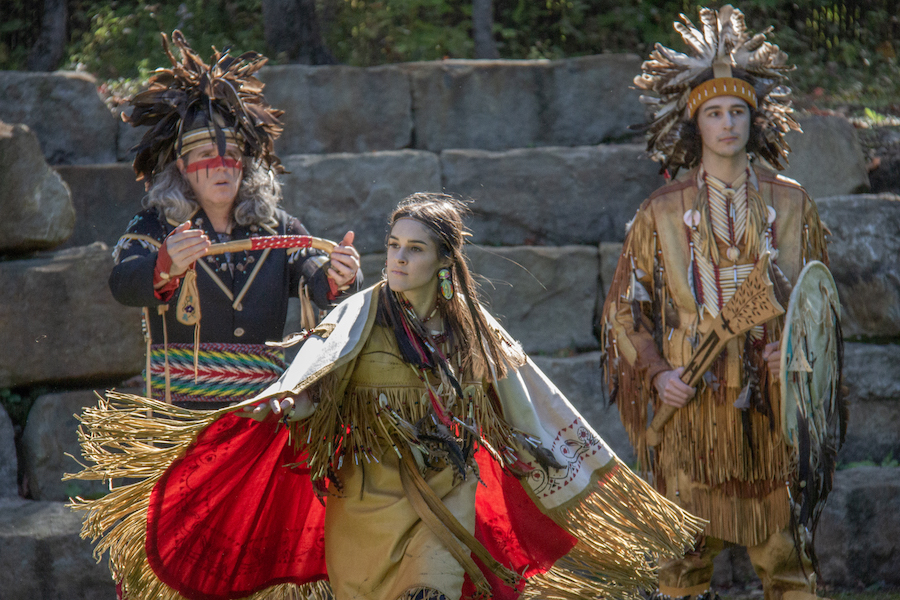 A woman dances in front of two men standing the background. All three figures are part of the First Nations Huron-Wendat tribe in Wendake, just outside of Quebec City. They are wearing traditional garb, including fringed animal hides and feathered headdresses. 