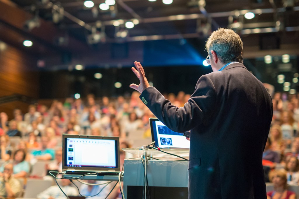 A view from behind the stage looking out at a conference speaker presenting to an audience of several hundred people as part of an event legacy project.