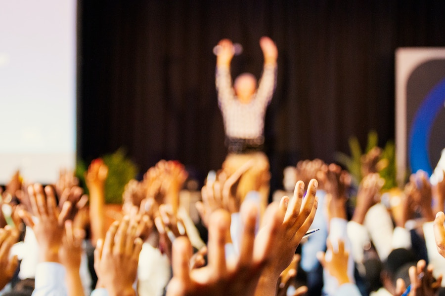 A view over a crowd sitting down at a business event, with just their raised hands visible in the camera frame. A blurry figure on stage has their hands up in the air.
