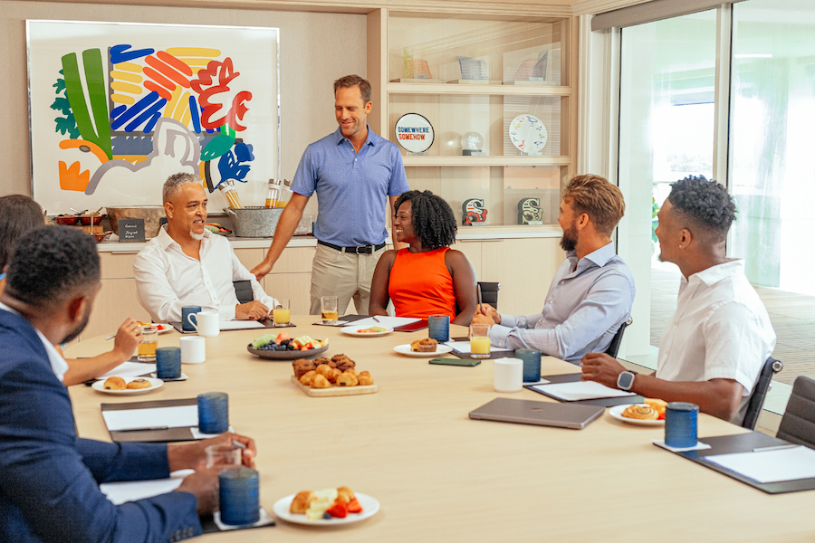 As part of an event theme around strength and resilience, a group of board member sit around a table with light refreshments in a brightly-lit room with colorful art and decor in the background.