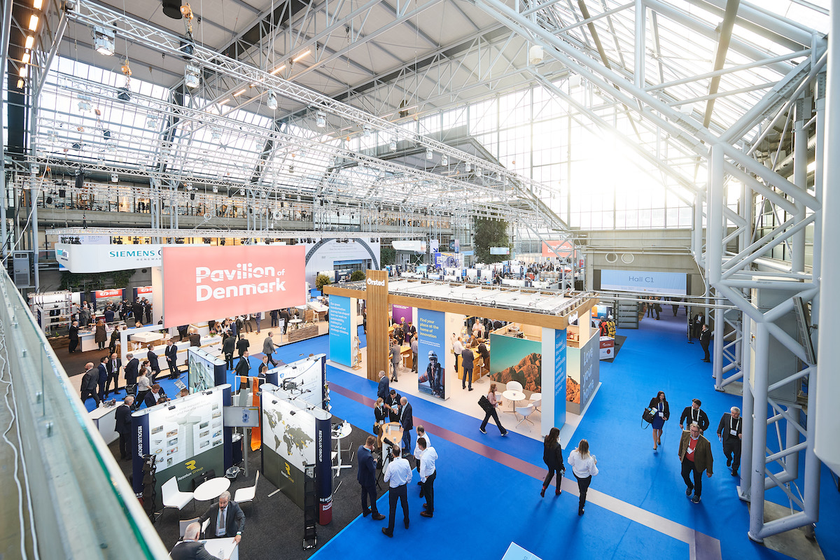 An interior view of a Danish congress, Electric City, held at a major convention center, with natural light streaming in from skylights above and a crowd of people walking around wind-energy-related booths below. This event served as an event legacy case study.