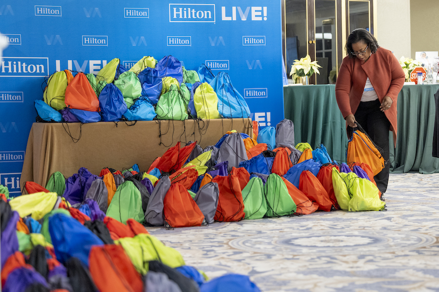 A woman bends over to place a bagged hygiene kit down among a pile of other bagged hygiene kits on a carpeted floor and on a table, with a large "Hilton Live!" wall-to-ceiling poster in the background. This kind of  kit-packing activity can be a great way to contribute to ESG goals for meetings.