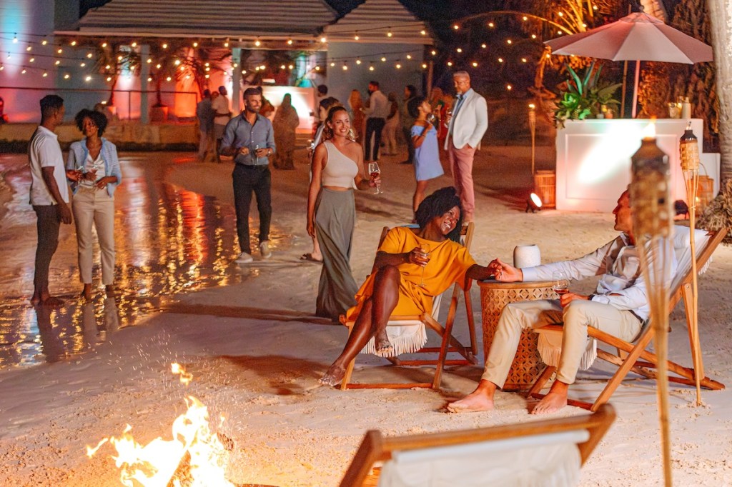 A group of about 20 to 30 people gather on the beach at night for a culture-themed evening event, with lounge chairs and a small bonfire.