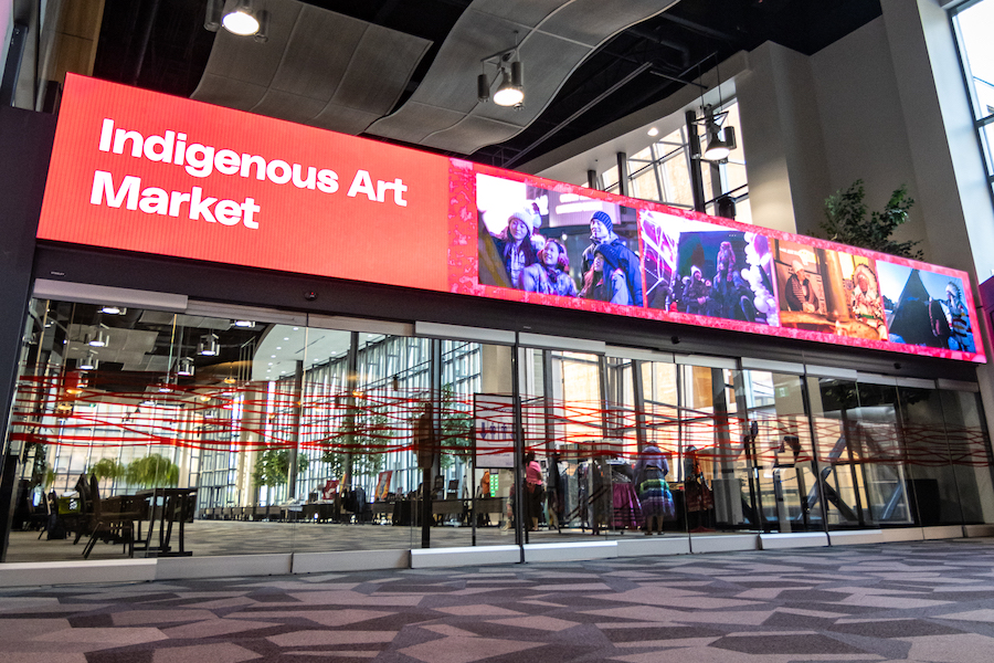 A large LED screen above the entrance to an exhibit hall in the Calgary TELUS Convention Center (CTCC) displays a sign for the Indigenous Art Market, with photos of Indigenous people displayed next to the event name and vendor booths visible beyond the glass doors below.