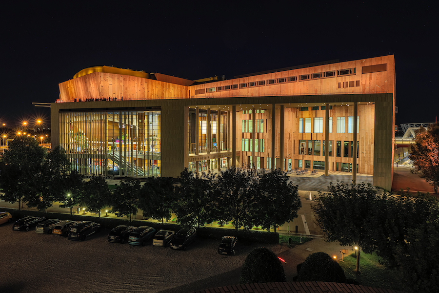 An exterior view of the MUPA cultural center at night. It is an ultra-modern building with a foyer encased in glass walls that are several storeys high and an interior siding of orange-gold tiles. The opportunities for cultural experiences abound at MUPA.