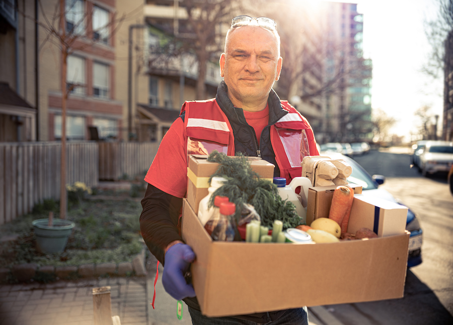 A man is seen standing on a sidewalk holding a cardboard box full of fresh produce, eggs, and other grocery goods. These kinds of food donation programs can be one way for events to give back and contribute to ESG goals.