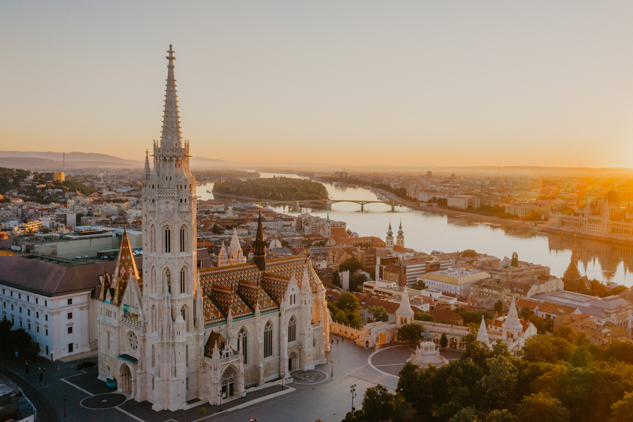 As aerial view of Matthias Church and the Danube shimmering behind it, with the waterfront serving as a favorite spot for visitors to enjoy cultural experiences. 