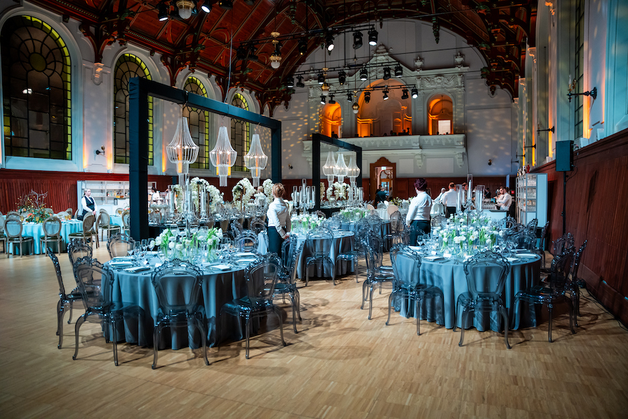An interior shot of the Royal Riding Hall in Budapest. An arched ceiling is flanked by large stained glass windows, and round tables decorated with flowers and candelabras fill the room. Hosting a gala event in a historical structure like this one turn a social gathering into a full cultural experience.