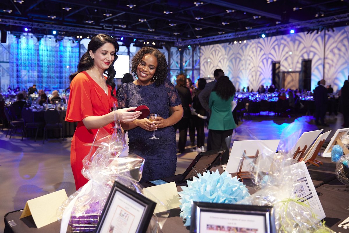 Two women stand side by side admiring event swag at the Calgary Telus Convention Center, with blue and purple lighting creating atmosphere in the background.