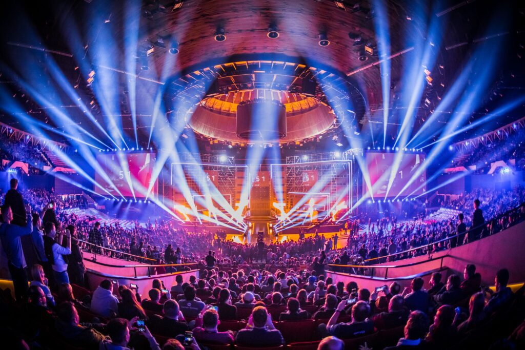 An e-sport tournament at the Katowice International Conference Centre in Poland. A crowd of approximately 500 people is seen batched in purple light facing a stage with multiple criss-crossing kinetic spotlights.