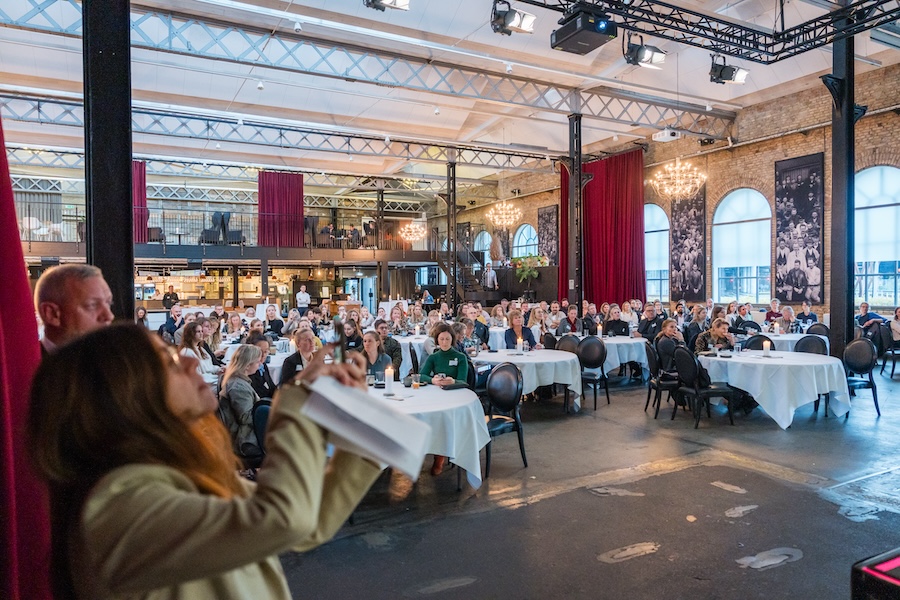 A view from the stage at a conference keynote address, with a woman on the left-hand side taking a photograph with her phone and a crowd of people seated at round dinner tables in the background. When it comes to measuring event legacy success, it's key to go beyond mere attendance numbers.