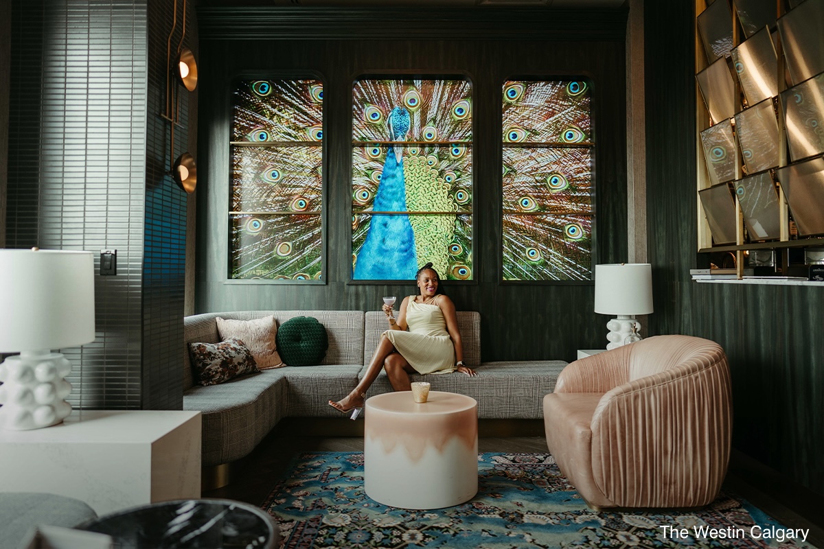 An interior shot of a lounge at the Westin Calgary, with a tiled series of paintings depicting a peacock on the back wall and a woman smiling and holding a drink amidst a set of cushy sofas as part of a bleisure experience at a business event.