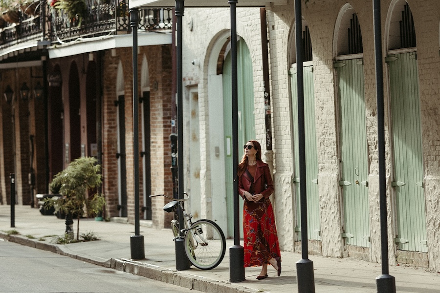 A woman leans against a thin black pillar that is supporting a balcony renovation on a heritage brick building in the French Quarter of New Orleans. Other brick buildings with elaborate filigree balcony railings can be seen in the background. Visiting historic neighborhoods is a great bleisure addition to business events and meetings.