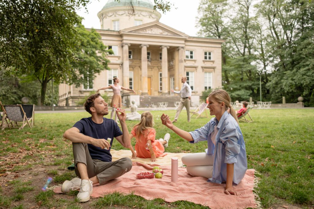 An idyllic scene outside a small palace in Warsaw, Poland, with a neoclassical design and four large columns along its entrance. A family of three is seen picnicking in the foreground, while two other adults practise ballet in the background.