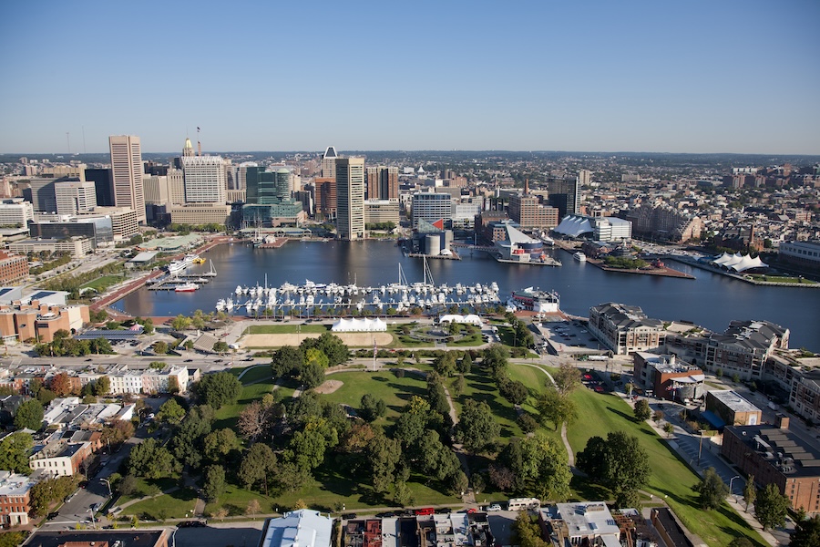 An aerial view of Baltimore's skyline, with a park in the foreground, an inlet of the harbor with docked boats, and the skyscrapers that are foundational to the city's tech hub in the distance.