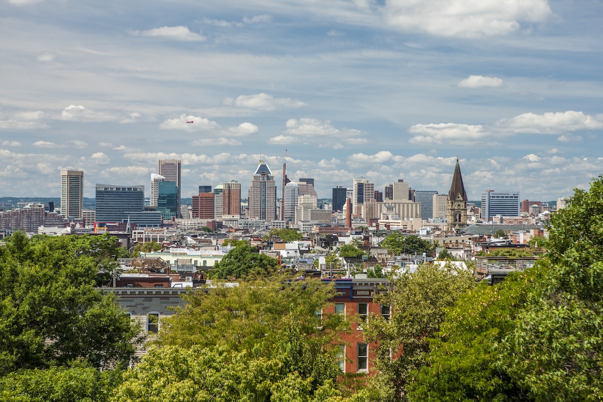 A skyline view of Baltimore, a city that's rapidly emerging as an up-and-coming tech hub ideal for hosting future-forward meetings and events. Green tree foliage is visible in the foreground, with low-rise Victorian brick buildings and a church tower in the middle ground and skyscrapers in the background.