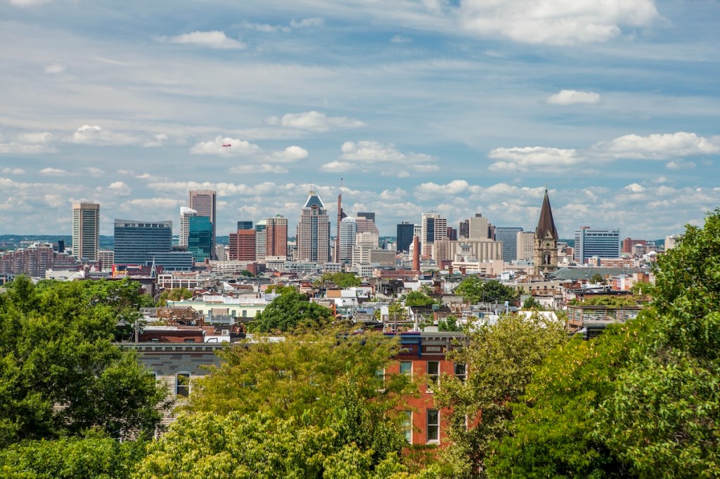 A skyline view of Baltimore, a city that's rapidly emerging as an up-and-coming tech hub ideal for hosting future-forward meetings and events. Green tree foliage is visible in the foreground, with low-rise Victorian brick buildings and a church tower in the middle ground and skyscrapers in the background.