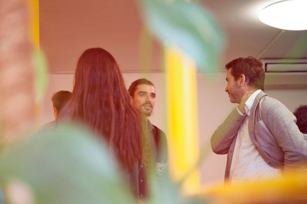 Three people are seen having a thoughtful discussion between leaves that are out of focus in the foreground. Two men are facing the camera, and a woman is seen from behind.