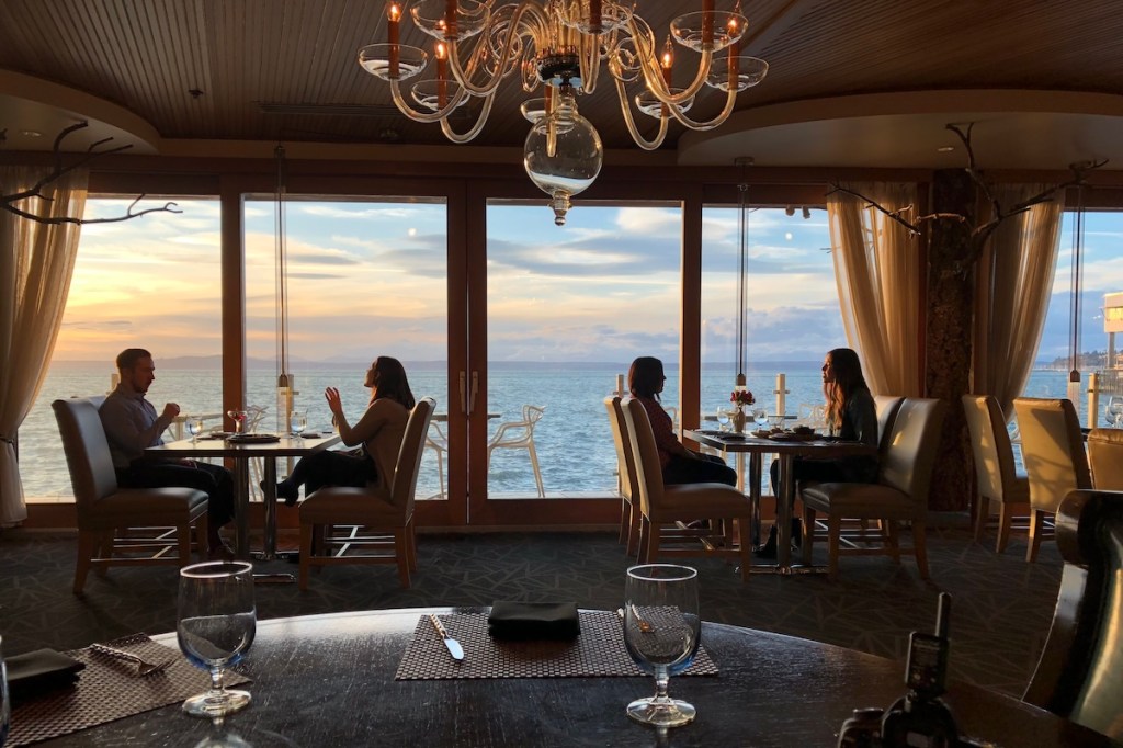 Two sets of people sit at round tables, all of them engaged in deep conversations. Just behind them is a large window facing out onto the ocean with an uninterrupted view of the water and the horizon. Intentional meetings should consider the type of setting that will be most conducive to meaningful networking.