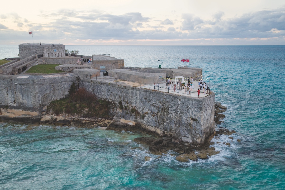 An aerial view of Bermuda's Keep Fort, a high-walled stone structure jutting out into the Atlantic ocean. A small group of executives mingle during an outdoor event on the platform, where they can enjoy a panoramic view of the clear aqua water as part of their corporate retreat's evening itinerary.