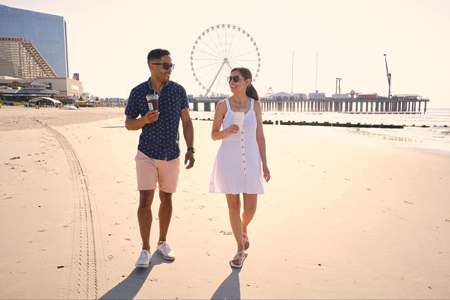Wearing summer attire and drinking iced coffees, a man and a woman walk side by side along one of Atlantic City's iconic beaches.