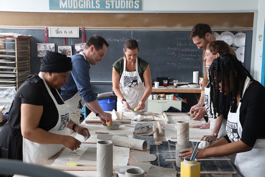 Four women and two men stand around a table where they are all leaning forward to work on ceramic projects as part of a MudGirl Studios workshop in Atlantic City.