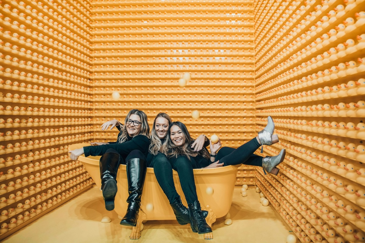 Three women sitting inside bathtub surrounded by rubber ducks