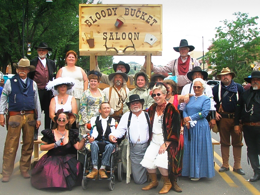 Approximately 20 people of mixed genders, ages, and abilities stand in three rows wearing authentic clothing from the pioneer days. The sign behind them reads, "Bloody Bucket Saloon."