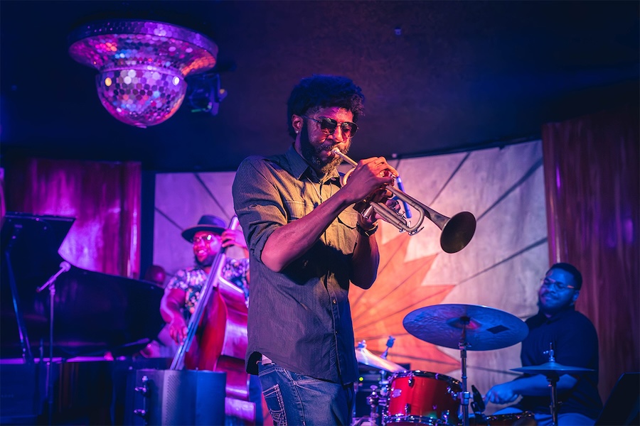 An interior shot of the live music stage at Cliff Bell's in Detroit. A man plays the trumpet in the foreground, with a bass player and a drum player in the background.