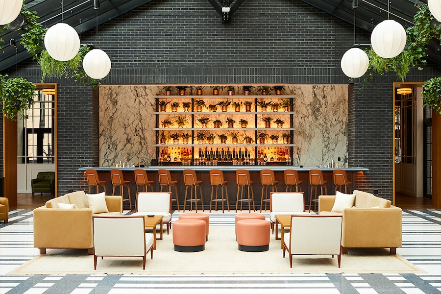 An interior shot of the Shinola Hotel in Detroit. Spherical lanterns hang from the ceiling, and the back wall features a sleek bar with a marble backing. The seating area consists of lounge chairs, coffee tables, and ottomans.