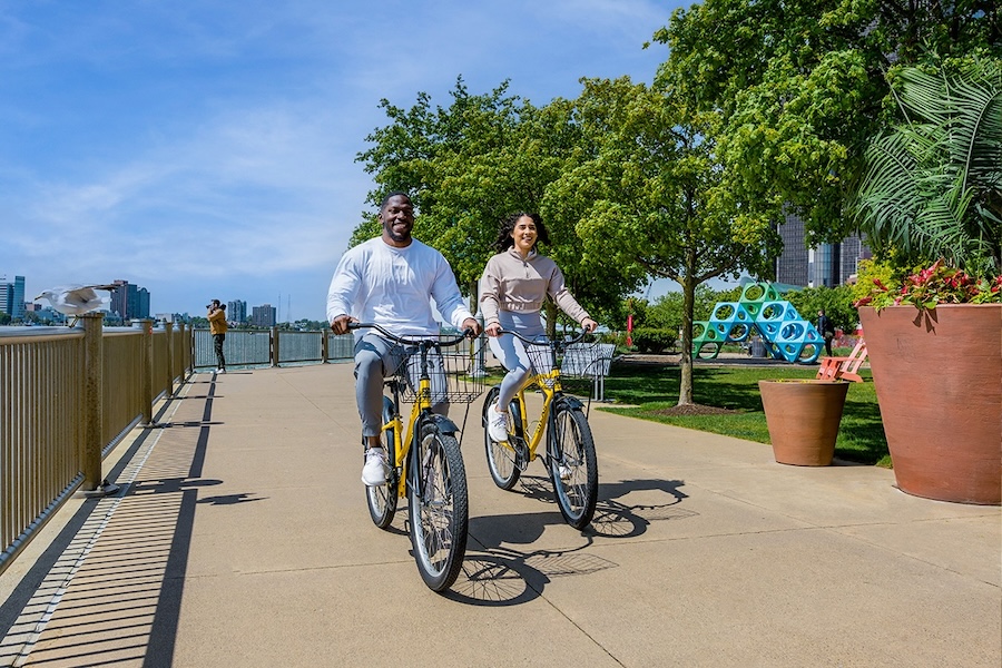 Two cyclists ride along one of Detroit's waterfront bike paths, with large planters, trees, and a modernist sculpture on the right-hand side.